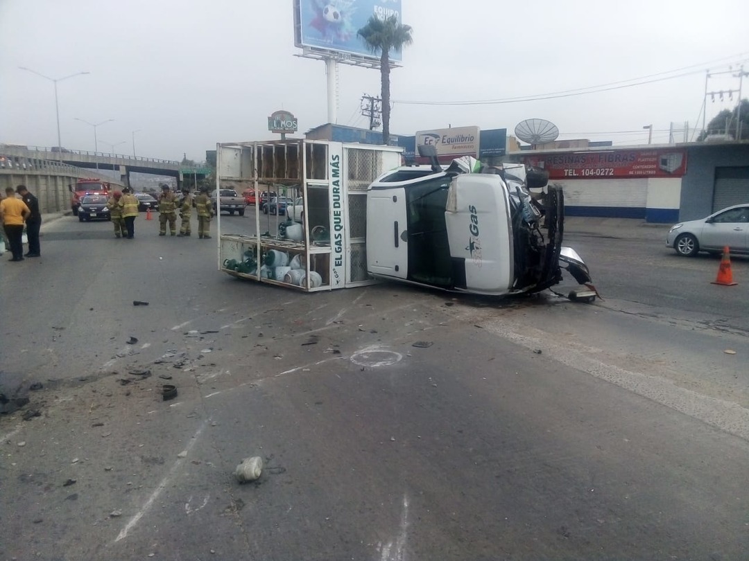 Vehículo volcado en la carretera junto a un quiosco de vidrio. Se observan escombros y personal de emergencias en la escena.