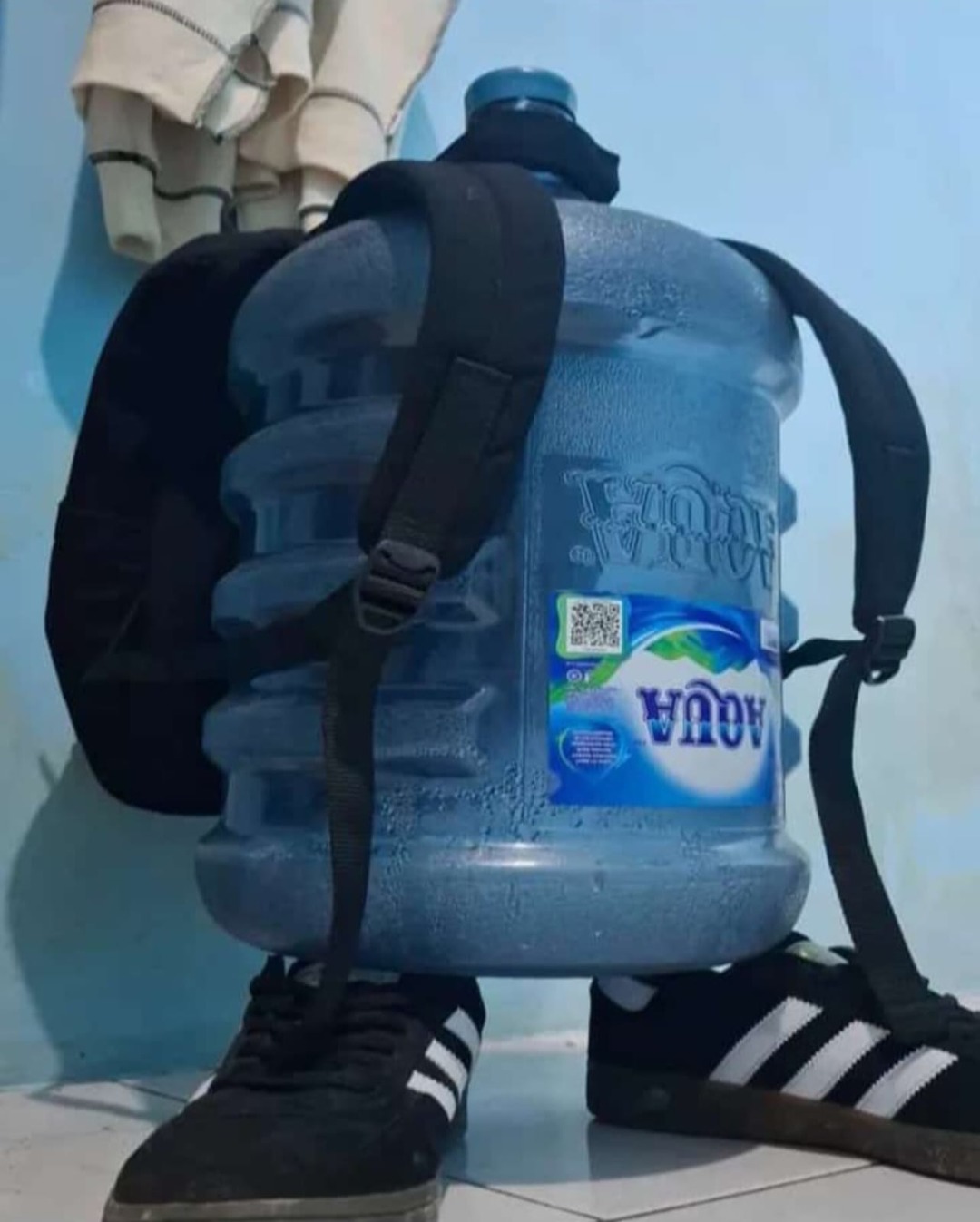 Mochila con tanque de agua de 5 galones, de color azul, con correas negras; hay una etiqueta azul que parece decir AQUA. En el suelo se ven tenis negros con marcas blancas. Sesión ID: 7847.