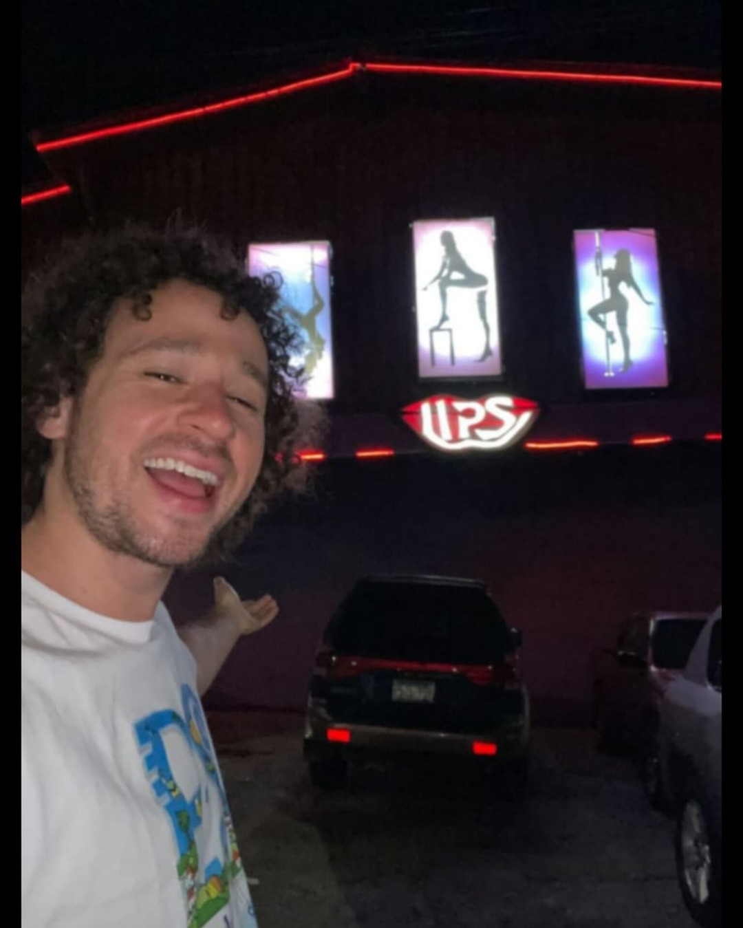 Selfie de un hombre sonriendo frente a un club con luces de neón y siluetas de baile en las pantallas.
