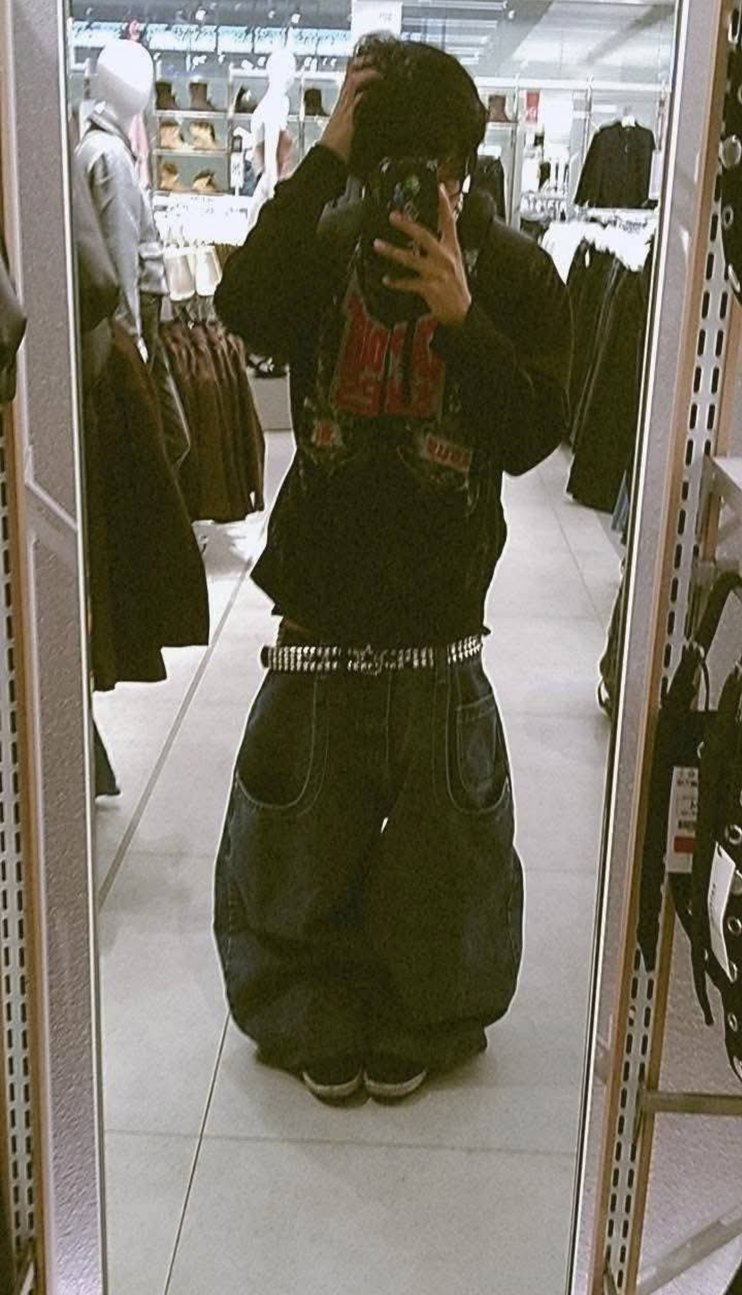 Persona tomando una selfie en una tienda de ropa. Lleva pantalones amplios tipo baggy con un cinturón plateado, sudadera negra con gráfico rojo y zapatones oscuros. El entorno parece una tienda con ropa colgada y percheros al fondo.