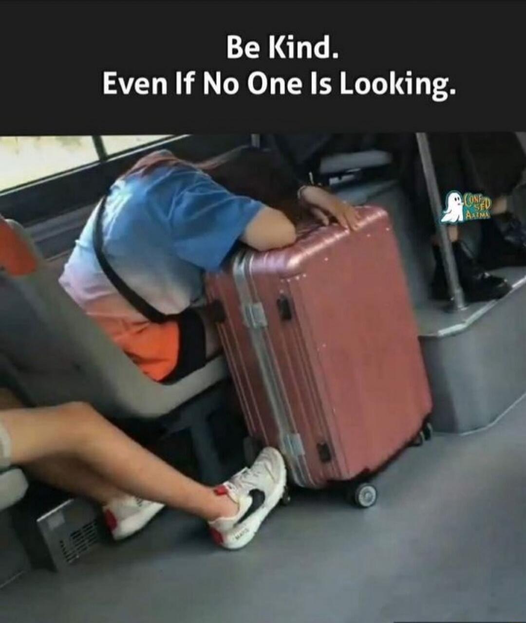 Be Kind. Even If No One Is Looking. A person rests on a bus seat with a pink suitcase beside them.