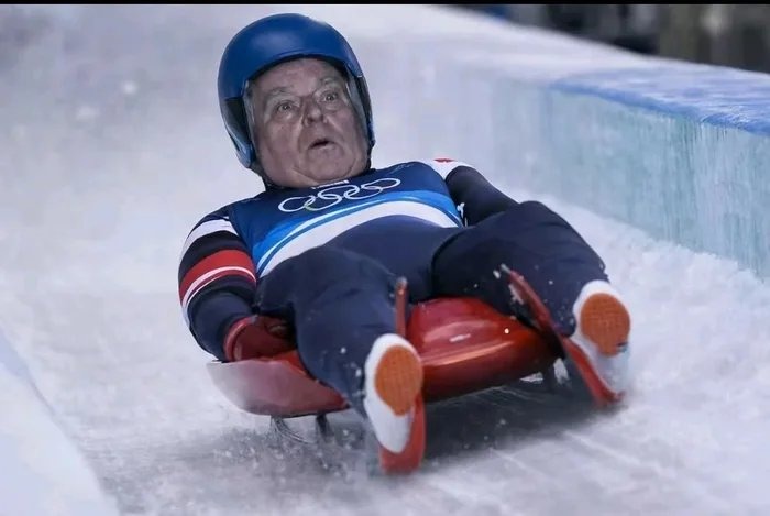 An older man wearing a blue helmet and a blue racing suit sits on a red sled, sliding down an icy track.