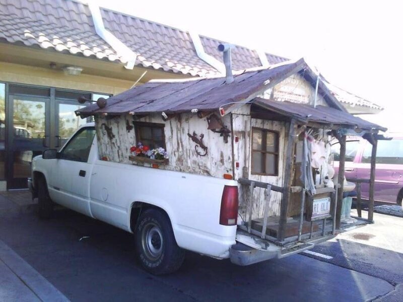 A white pickup truck has a shabby wooden shack built on its bed, with a tarp roof and small windows, parked in a driveway.