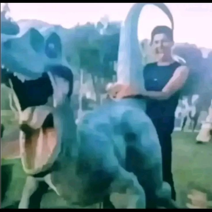 Una persona posa junto a una estatua de dinosaurio azul en un parque.
