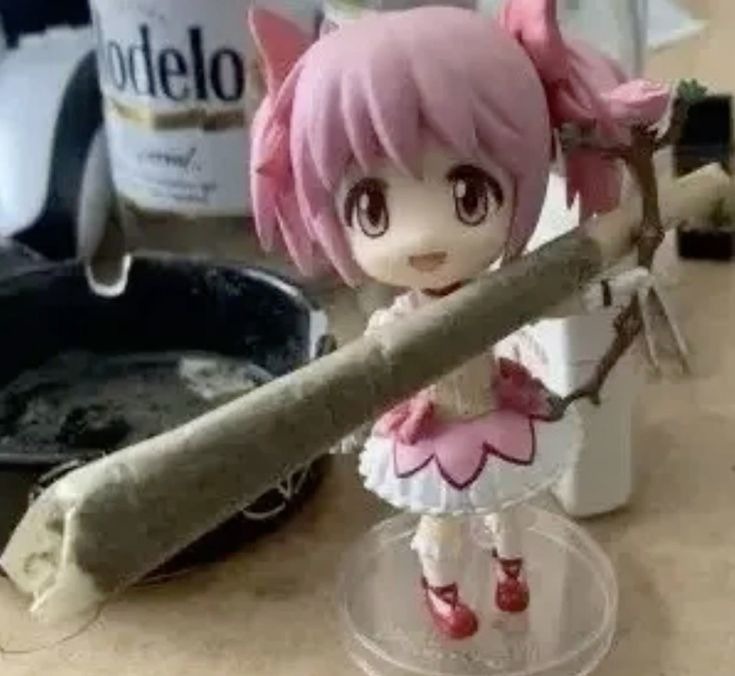 No speech detected. The image shows a figurine of a character with pink hair and a pink dress, holding a large joint. In the background, there is a bottle of 
