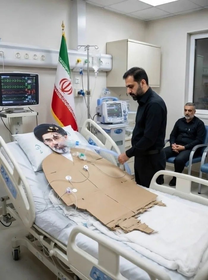 The image shows a hospital bed with a cardboard cutout of a person lying in it, dressed in religious attire. A man in a black shirt is attending to the cutout, holding a ventilator tube. Another man sits in a chair in the background. An Iranian flag is visible on a pole next to the bed. Medical equipment, including a monitor displaying vital signs,