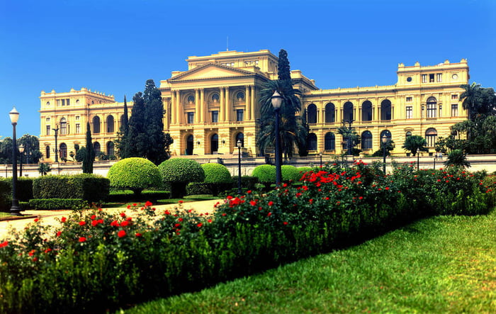 Museu do Ipiranga. Edifício histórico com arquitetura clássica, jardim bem cuidado com flores vermelhas, arbustos verdes e gramado, sob um céu azul claro.