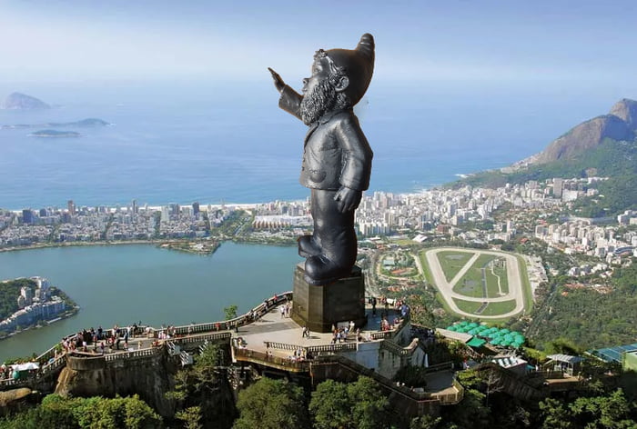 Uma estátua de um gnomo com barba e chapéu pontudo, substituindo o Cristo Redentor, com vista para a cidade do Rio de Janeiro, a lagoa e o oceano.