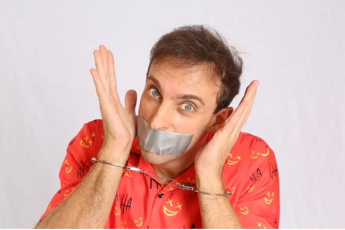 HA HA (texto na camisa). Um homem com fita adesiva na boca e algemas nos pulsos, com as mãos levantadas e uma expressão de surpresa ou desespero.