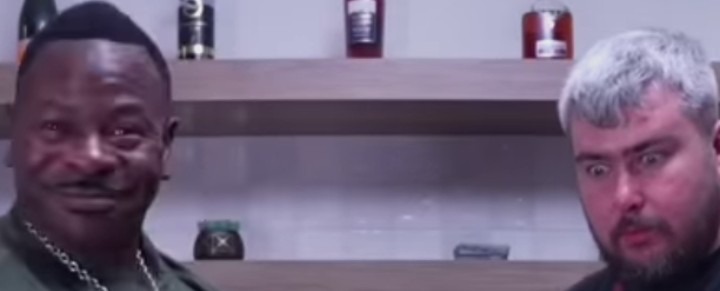 Dos hombres, uno sonriendo con bigote y el otro con una expresión de sorpresa o asombro, mirando algo fuera de cámara. Hay botellas en estantes al fondo.