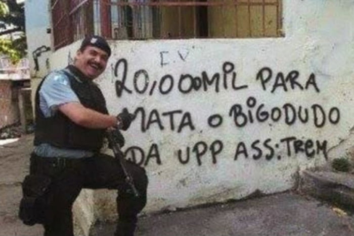 20.000 MIL PARA MATA O BIGODUDO DA UPP ASS: TREM. Um policial sorridente, aparentemente o 'bigodudo' mencionado, posa em frente à pichação que oferece uma recompensa pela sua morte.