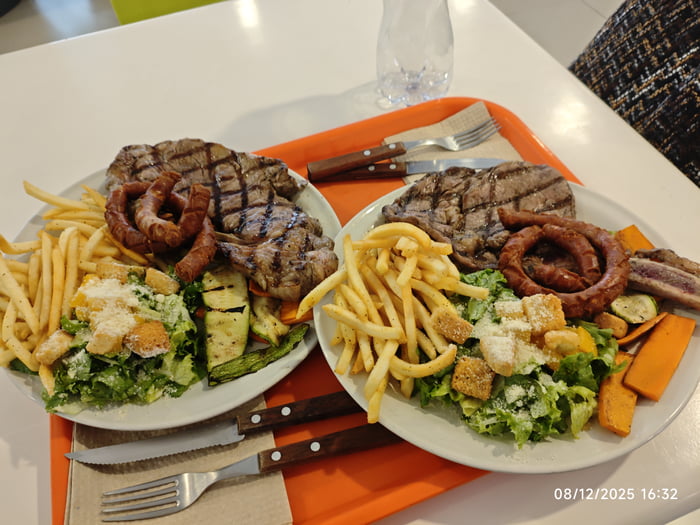 Texto en pantalla: 08/12/2025 16:32. Dos platos grandes de comida con bistec a la parrilla, salchichas, patatas fritas, ensalada César con picatostes y queso, y verduras asadas como calabacín y zanahoria.