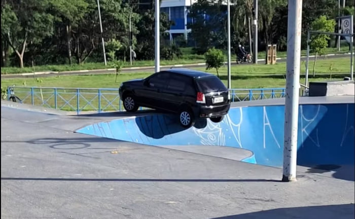 Um carro preto está preso em uma pista de skate, com as rodas traseiras suspensas no ar sobre o bowl. O veículo parece estar em uma situação inusitada e cômica.