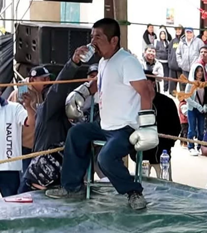 Un hombre con guantes de boxeo está sentado en una silla en un ring, bebiendo de una lata. Hay espectadores alrededor.