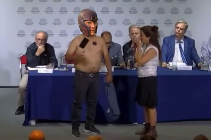 Un hombre sin camisa con un casco de Magneto en la cabeza y un teléfono pegado al pecho, de pie en un evento formal. Una mujer está a su lado. La escena parece un meme o un momento viral.