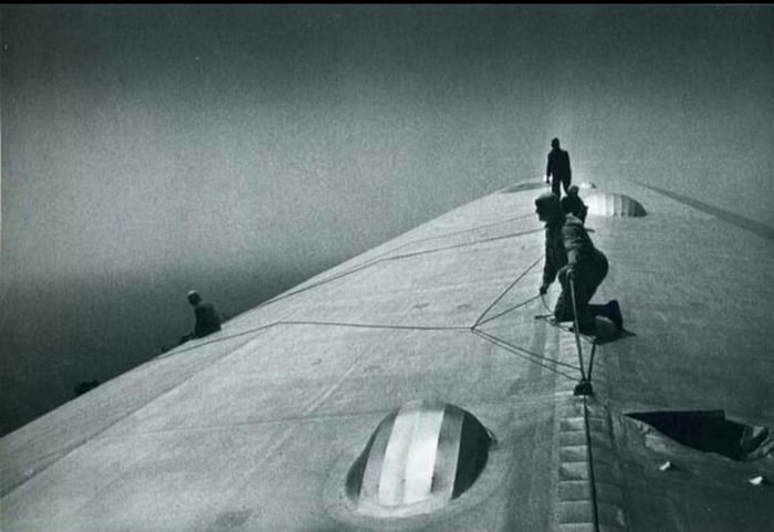 Pessoas trabalhando no topo de um dirigível ou zepelim, possivelmente realizando manutenção ou inspeção. A imagem é em preto e branco, sugerindo uma época histórica.