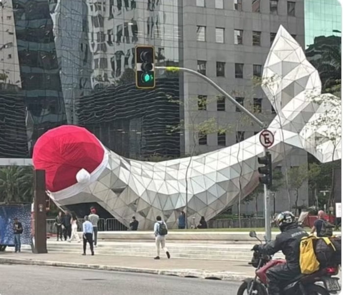 Uma grande estrutura geométrica moderna, que se assemelha à cauda de uma baleia, está decorada com um enorme chapéu de Papai Noel vermelho. Pessoas caminham nas proximidades. Um semáforo está verde. Um motociclista é visível. Sinal de trânsito com 'E' cortado.
