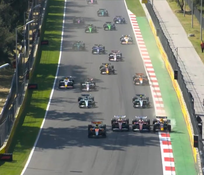 Una vista aérea de numerosos coches de Fórmula 1 en una pista de carreras, posiblemente al inicio de una carrera o durante una vuelta de formación. Los coches están alineados en varias filas, ocupando gran parte del ancho de la pista. Se pueden ver zonas verdes y bordillos a rayas rojas y blancas a los lados de la pista.