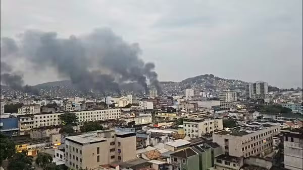 Fumaça escura e densa subindo sobre uma área urbana, com colinas ao fundo densamente povoadas por edifícios. Múltiplos focos de fumaça são visíveis, indicando um evento significativo, possivelmente incêndios.