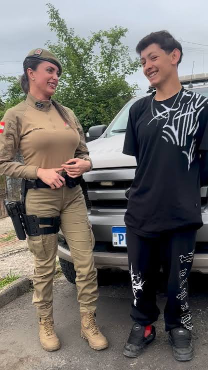 Uma mulher em uniforme militar ou policial e um jovem sorrindo, em pé ao lado de um veículo. A mulher tem o nome 'ROCHELI' visível em seu uniforme.