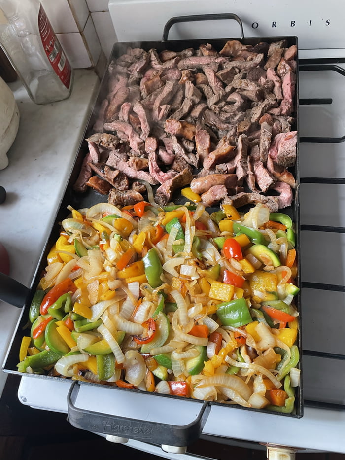 Carne en tiras y verduras salteadas (cebolla, pimientos de varios colores, calabacín) cocinándose en una plancha grande sobre una estufa. Se puede leer 'ORBIS' en la estufa y 'La Planchita' en la plancha.