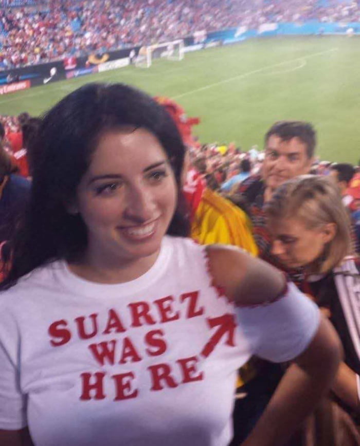 Uma mulher sorridente em um estádio de futebol, vestindo uma camiseta branca com a frase 'SUAREZ WAS HERE' em vermelho. Seu ombro direito tem uma marca que simula uma mordida, fazendo referência ao jogador Luis Suárez.