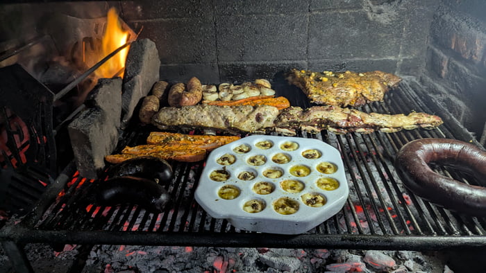 Una parrilla llena de diferentes tipos de carne, incluyendo costillas, chorizos y morcillas, cocinándose sobre brasas ardientes. También hay una bandeja con porciones individuales, posiblemente queso derretido o algún aperitivo, en la parrilla. El fuego es visible a la izquierda.