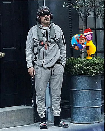 Un hombre con bigote, gafas de sol y auriculares, vestido con un chándal gris, de pie frente a una entrada. Sostiene lo que parece ser un cigarrillo. A su derecha, hay una imagen editada con un personaje similar a Mario y otro personaje amarillo con un gorro de Papá Noel.