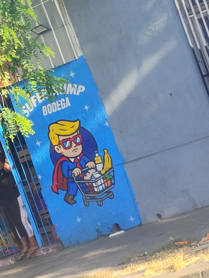 Mural en una pared azul con estrellas blancas que dice 'SUPER TRUMP BODEGA'. Muestra un personaje de dibujos animados con cabello rubio, gafas de sol, traje, corbata roja y una capa roja, que se asemeja a Donald Trump. El personaje empuja un carrito de compras lleno de comestibles como leche, plátanos y una botella, con una expresión seria.