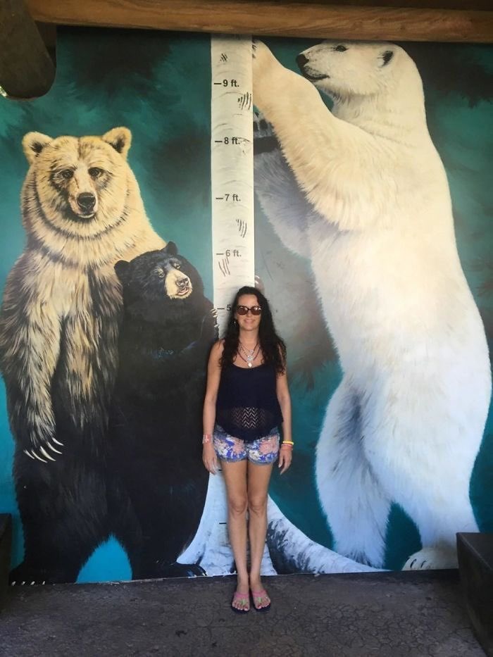 A woman stands in front of a mural depicting a grizzly bear, a black bear, and a polar bear, with a height chart showing measurements in feet: -9 ft., -8 ft., -7 ft., -6 ft.