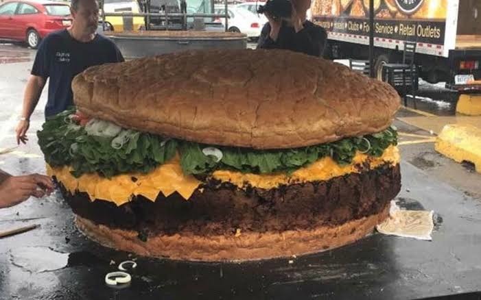 Una hamburguesa gigante con pan, carne, queso, lechuga y cebolla. Varias personas están alrededor de la enorme hamburguesa.