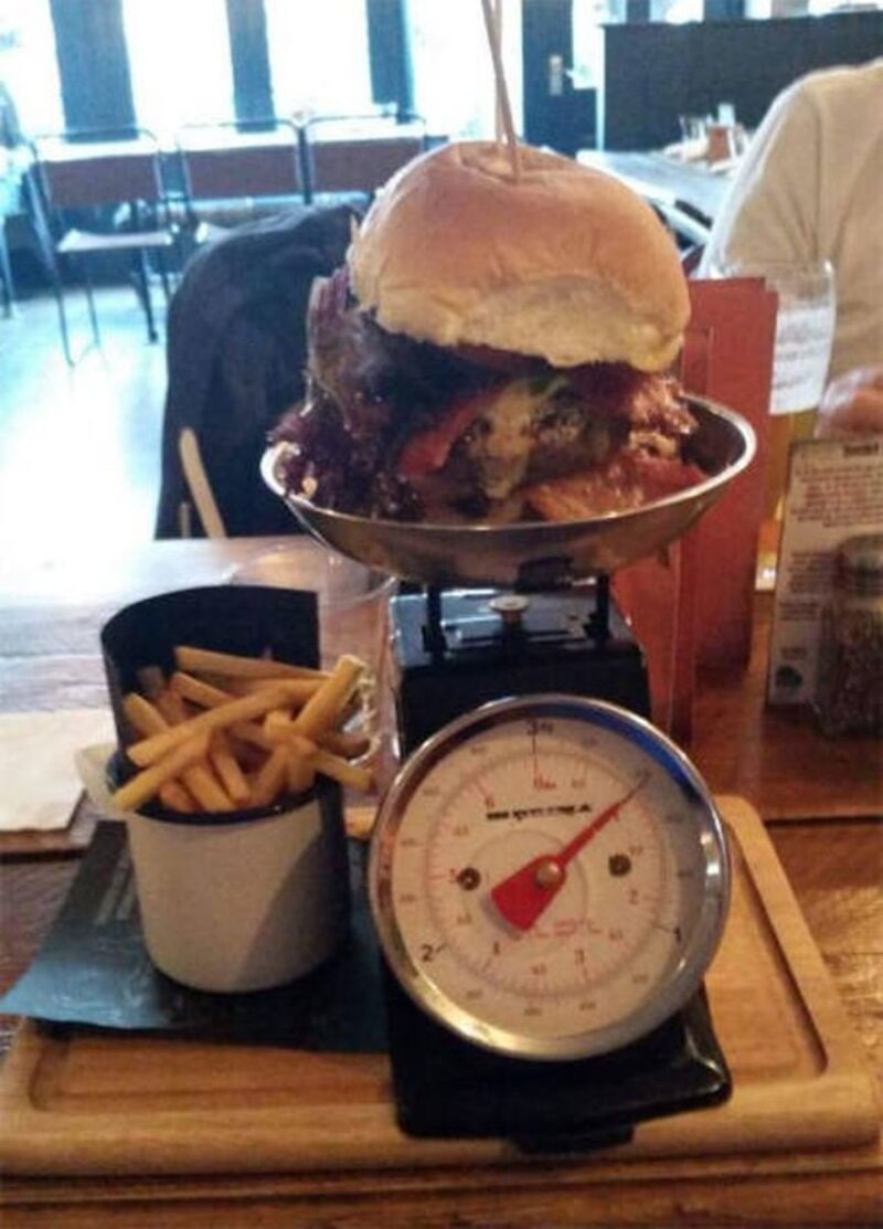 A massive burger is served on a kitchen scale, indicating its huge weight. Next to it is a cup of french fries. The scale's dial shows a significant measurement, highlighting the burger's enormous size. The setting appears to be a restaurant.