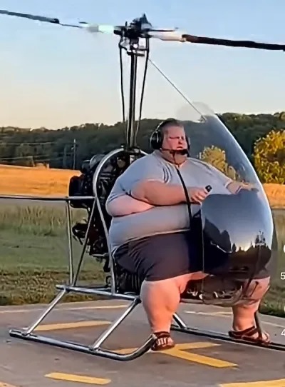 A very overweight person is sitting in a small, open-cockpit helicopter or gyrocopter, wearing a headset, seemingly preparing for a flight. The person's size appears to be a tight fit for the aircraft.