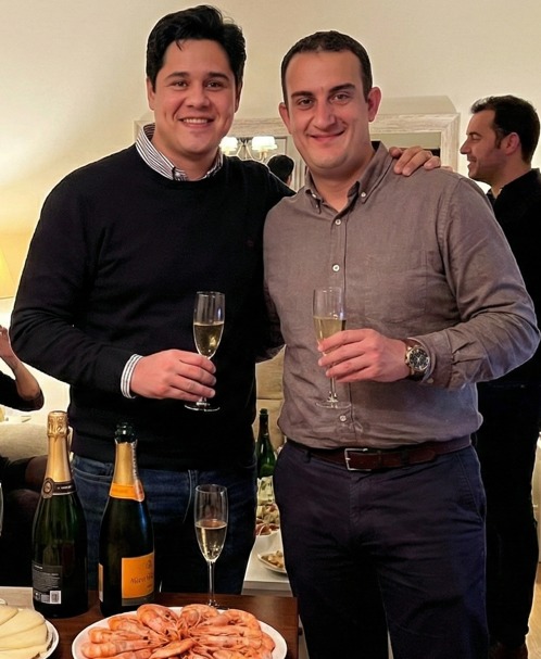 Dos hombres sonriendo y brindando con copas de champán en una reunión social. Hay botellas de champán, gambas y queso en la mesa.