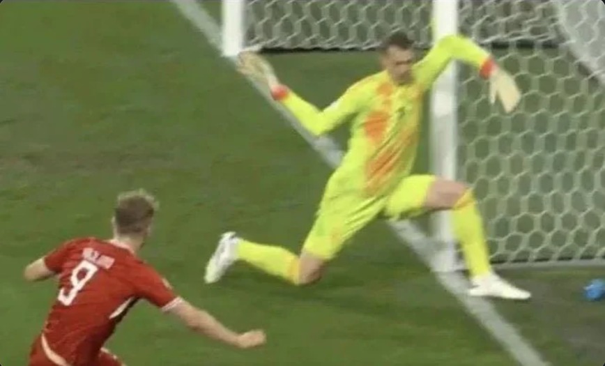 Um goleiro em um uniforme amarelo-limão está em uma pose desajeitada e acrobática perto da baliza, enquanto um jogador de camisa vermelha com o número 9 está por perto no campo de futebol.