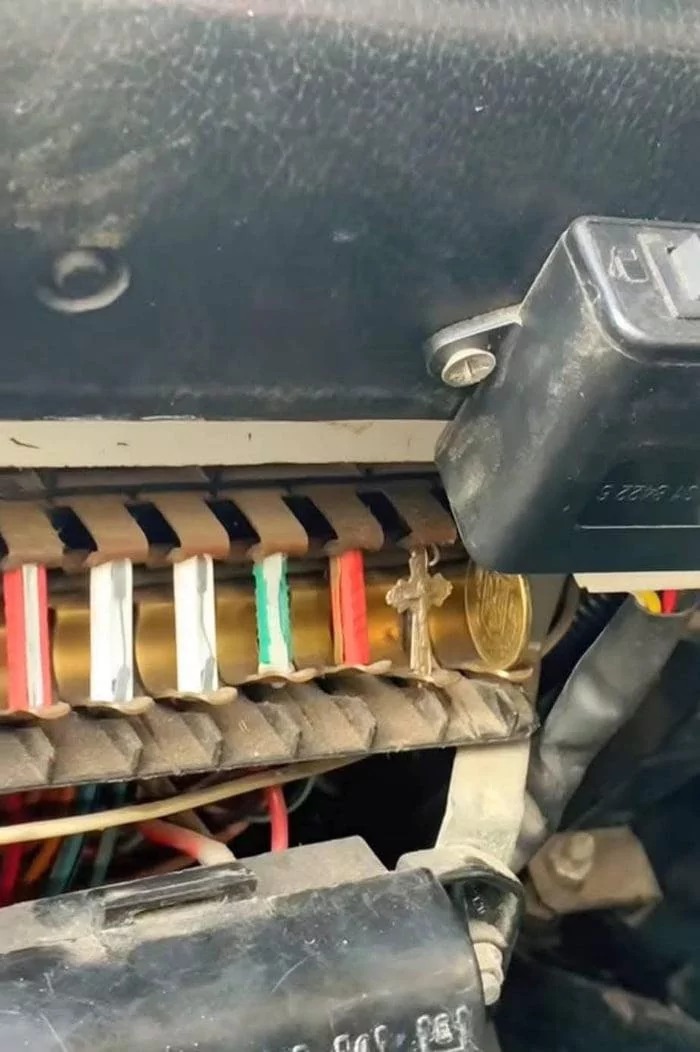 A close-up of a car's fuse box where standard fuses have been replaced with improvised items such as wires wrapped in tape, a small cross pendant, and a coin. This is a dangerous and improper electrical repair.