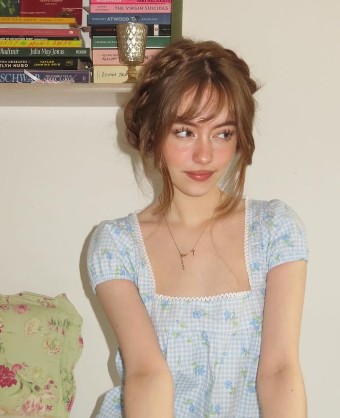 A young woman with braided hair and bangs, wearing a light blue gingham dress with small floral patterns and white lace trim. She is looking to the side with a gentle expression. A bookshelf with various books is visible in the background.