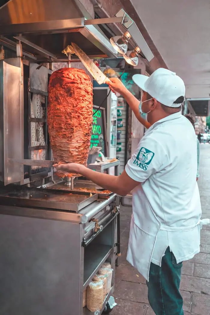 Un cocinero preparando tacos al pastor o shawarma de un gran asador vertical de carne. El cocinero lleva una gorra, mascarilla y un uniforme con el logo de IMSS. Se ven tortillas en bolsas en un estante debajo.
