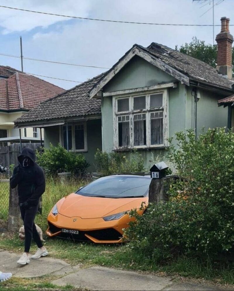 An orange Lamborghini Huracan is parked on a grassy patch next to a dilapidated, old house. A person in a black hoodie and mask walks past with a small dog. The house has peeling paint and overgrown bushes. A mailbox with the number 16 is visible. The car's license plate reads ZH-1023.