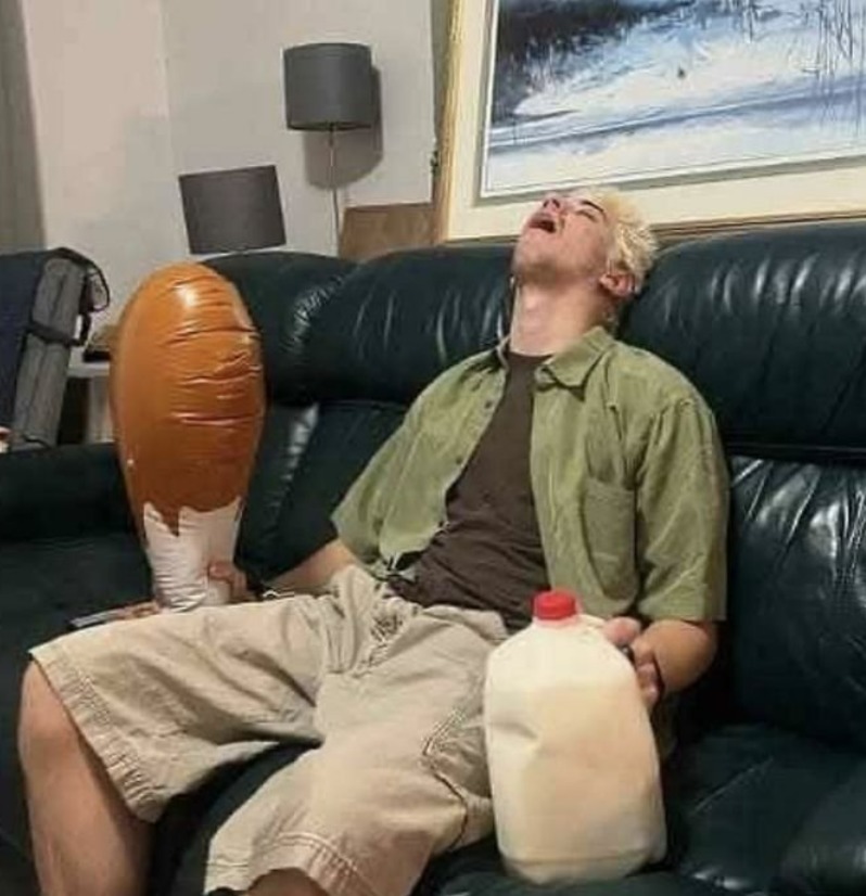 Un hombre joven con cabello rubio platino está sentado en un sofá de cuero, con la cabeza echada hacia atrás y la boca abierta en una expresión de éxtasis o agotamiento. Sostiene una jarra de leche en una mano y a su lado hay un objeto inflable con forma de cono de helado.