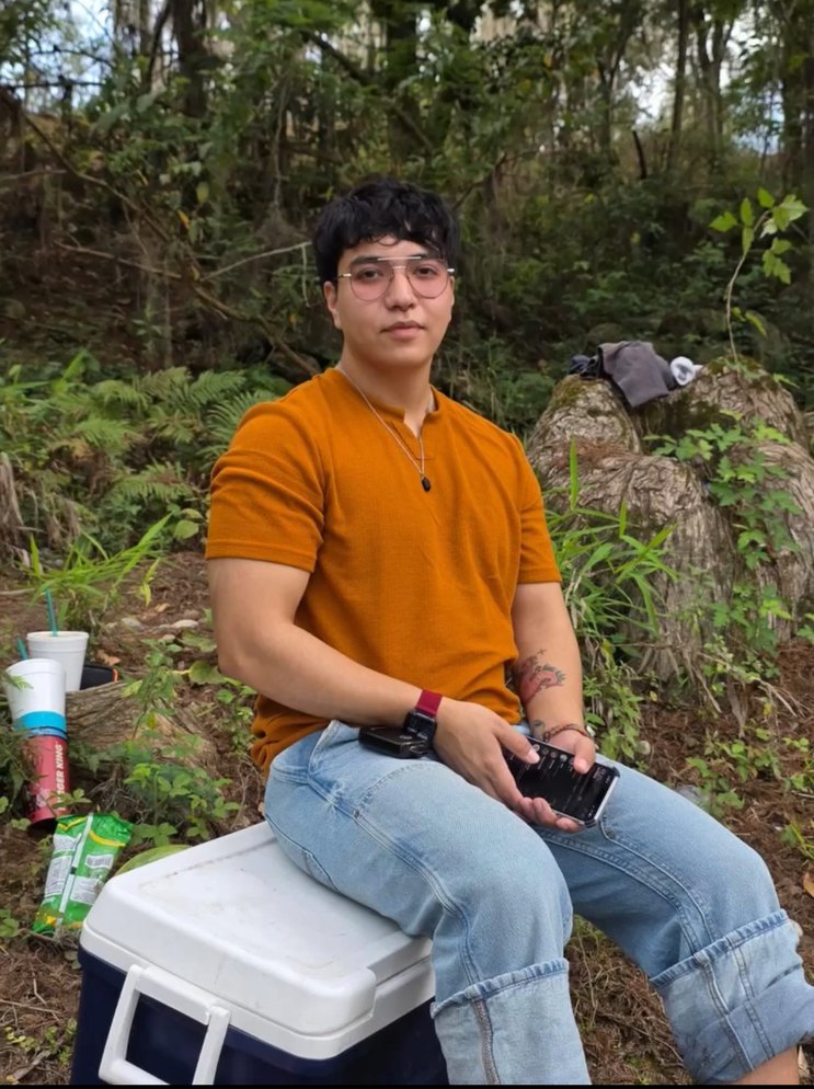Un joven está sentado en un refrigerador portátil al aire libre, rodeado de vegetación.