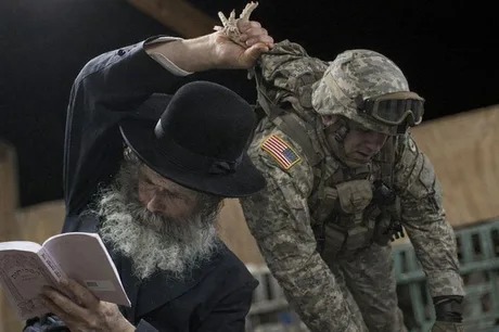 An older man with a long grey beard, wearing a black hat and coat, is reading a book. His arm is raised, holding onto the arm of a soldier in camouflage uniform and helmet. The soldier is bending over, possibly engaged in a task.