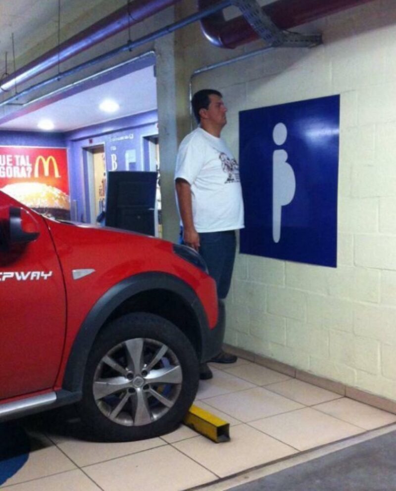 A man with a large belly stands next to a blue sign depicting a pregnant person, humorously comparing himself to the image. In the background, a McDonald's advertisement reads 