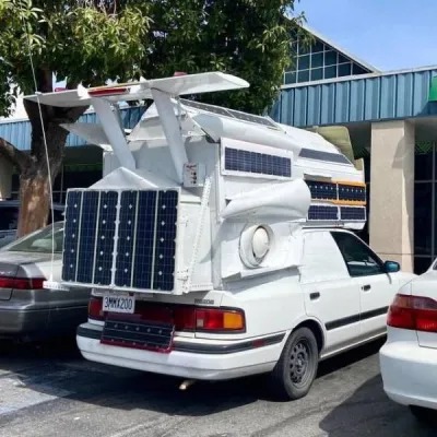 A white car heavily modified with airplane parts and numerous solar panels, parked in a lot. The modifications include wings, vents, and panels covering the roof and rear of the vehicle.