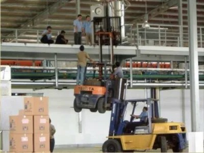 Workers are using two forklifts in an extremely dangerous and unsafe manner to reach a higher platform. One forklift is lifting another forklift, with a person standing on the elevated forklift's platform, while other workers observe from above.