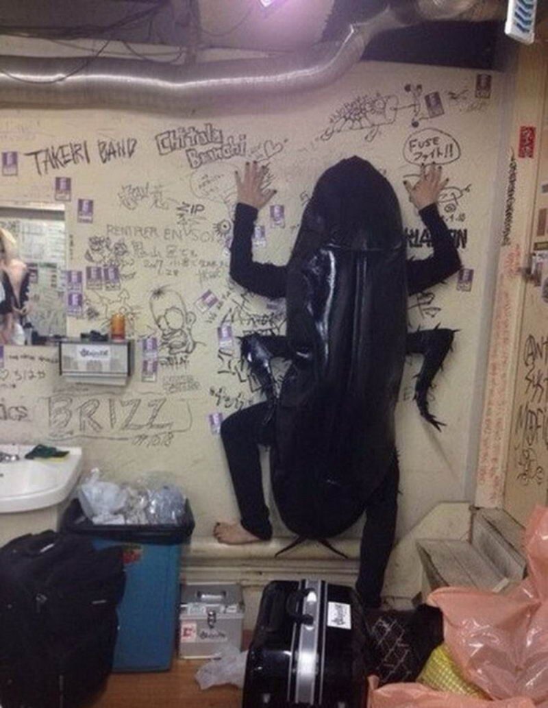 A person in a large black cockroach costume is standing against a graffiti-covered wall. Text on the wall includes 'TAKERI BAND', 'Chitola Brunch', 'Fuse Fight!!', 'ZIP', 'BRIZZ', and a date '07.10.18'. The room appears to be a messy backstage area with a sink, trash can, and luggage.