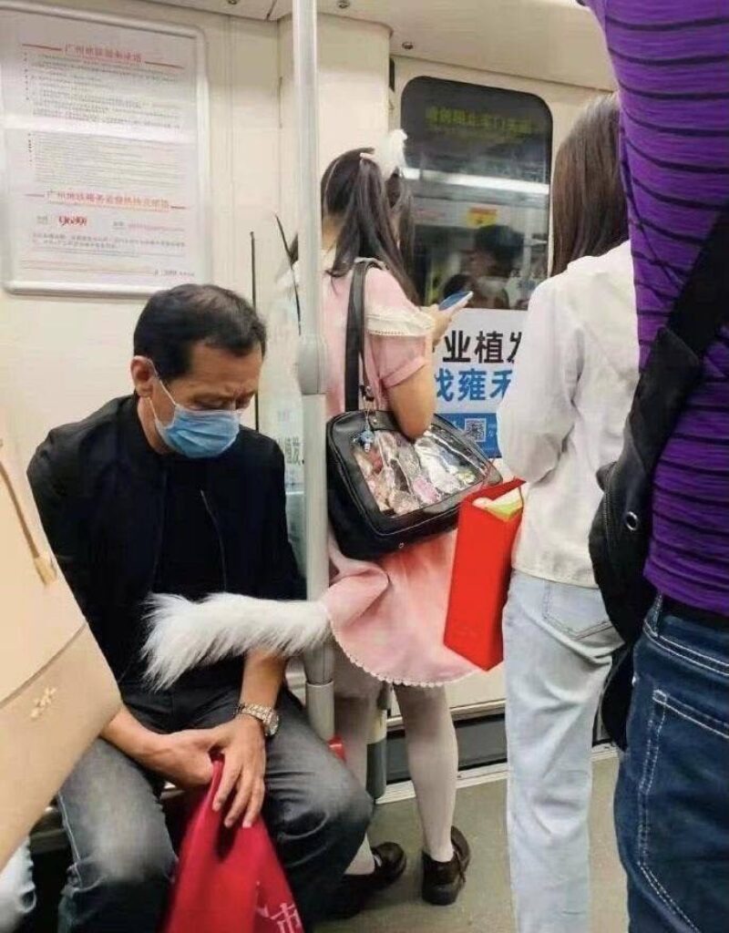 A man wearing a face mask is sitting on a subway train. Next to him, a person in a pink dress, white tights, and a white furry tail is standing, looking at their phone. The person also has cat ears. Other passengers are visible in the background. Chinese text is visible on signs and advertisements inside the subway car, including 