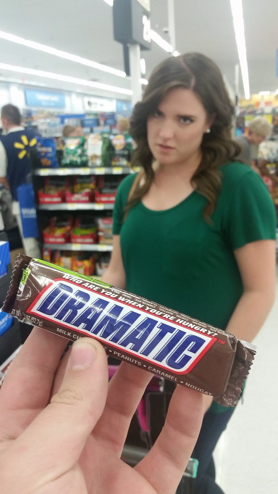 DRAMATIC WHO ARE YOU WHEN YOU'RE HUNGRY? MILK CHOCOLATE PEANUTS CARAMEL NOUGAT. A person holds a Snickers bar with 'DRAMATIC' written on it in front of a woman with a dramatic or annoyed expression in a supermarket.