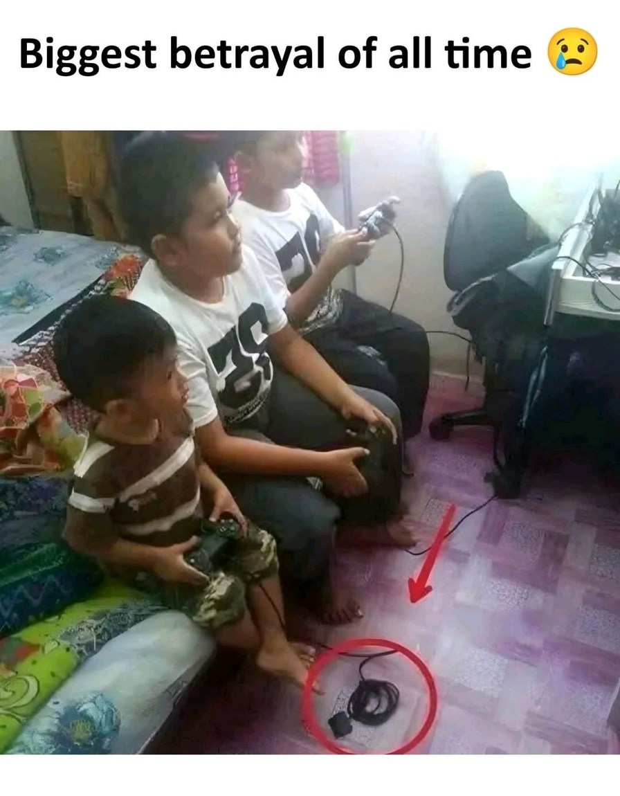 Biggest betrayal of all time 😭. Three boys are playing video games. The youngest boy is holding a controller, but his controller's cable is unplugged and lying on the floor, circled in red, meaning he's not actually playing.