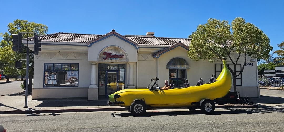 A man is driving a custom-built car shaped like a giant banana, parked in front of a Fosters Freeze restaurant. The building has the address 1593 and signs for 'DAILY' and 'CHICKEN BOSS'.